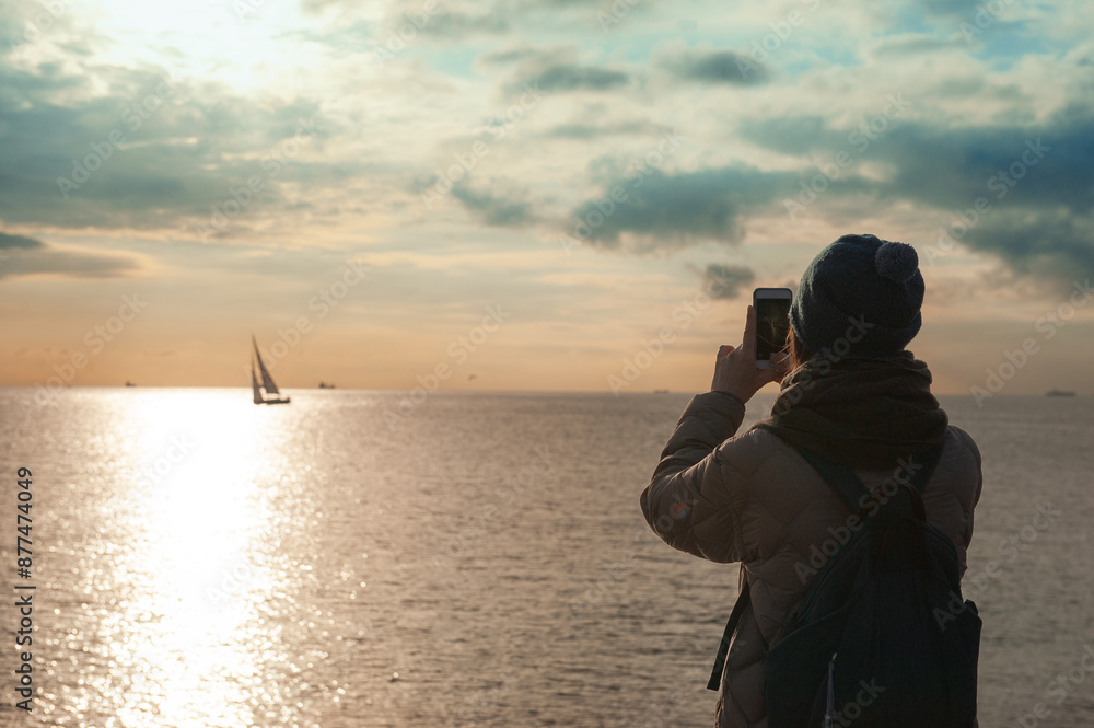 Woman in a winter coat and beanie takes a photo of the sunset over the sea, capturing a sailboat on the horizon Woman in a winter coat and beanie takes a photo of the sunset over the sea, capturing a sailboat on the horizon
