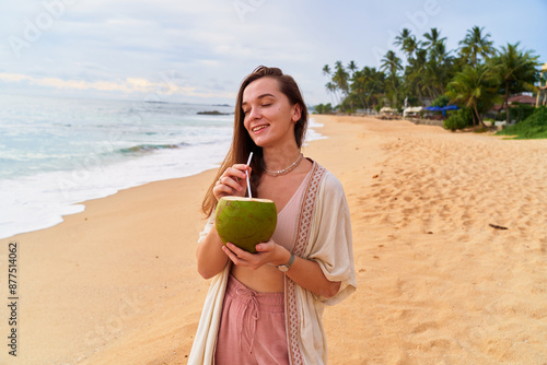 Young boho girl traveler holds in hands a tropical green coconut with a straw on ocean shore photo