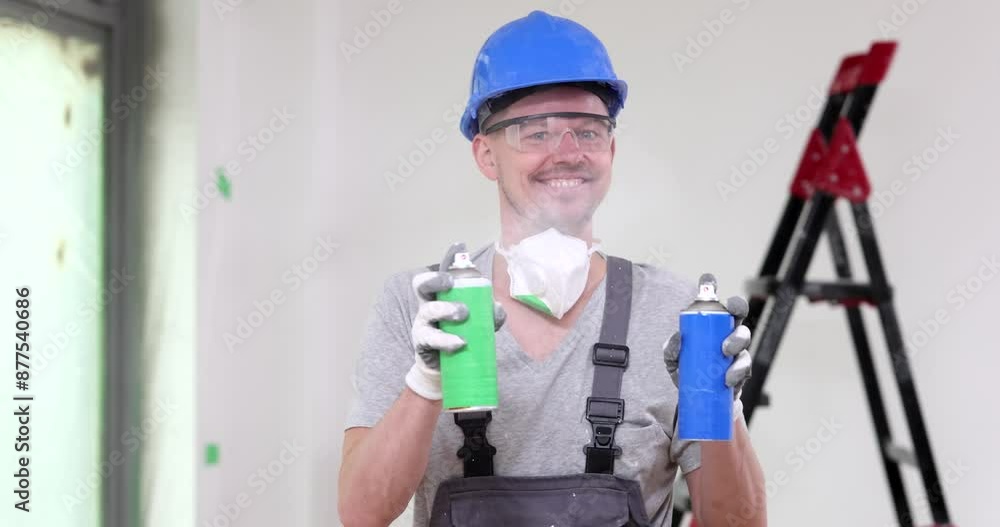 Happy foreman sprays white paint from green and blue bottles. Cheerful man in protective helmet and goggles smiles looking in camera slow motion Happy foreman sprays white paint from green and blue bottles. Cheerful man in protective helmet and goggles smiles looking in camera slow motion