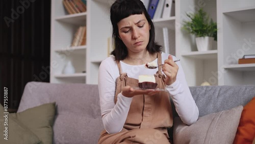 Portrait of sad young woman dealing with stress by eating food sitting on sofa at home Upset female eating cake indoors alone Mental heath problem