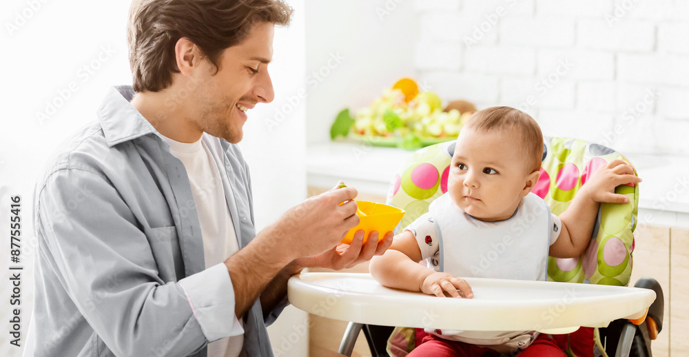 A father is feeding his baby in a high chair in a kitchen. The baby is wearing a bib and sitting in a brightly colored high chair. The father is smiling and looking at his baby. A father is feeding his baby in a high chair in a kitchen. The baby is wearing a bib and sitting in a brightly colored high chair. The father is smiling and looking at his baby.