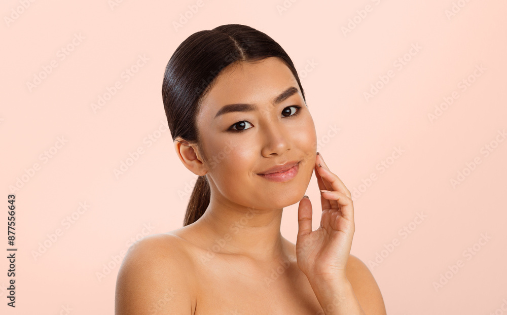 Asian young woman with long, black hair pulled back in a ponytail, touching her cheek with a gentle hand. She is smiling softly and has a natural, radiant complexion. Asian young woman with long, black hair pulled back in a ponytail, touching her cheek with a gentle hand. She is smiling softly and has a natural, radiant complexion.