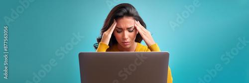 A young woman is seated behind her laptop, grasping her head in distress, positioned against a backdrop of light blue with ample space reserved for text.