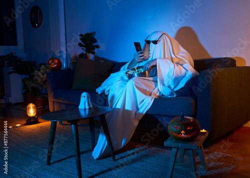 Halloween scary movie night. Person in ghost costume with sunglasses and pumpkin decor texting and eating popcorn while watching tv in sofa