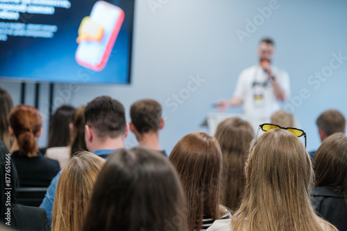 Audience attentively listening to a speaker at a business conference. The presenter speaks while attendees focus on the presentation screen.