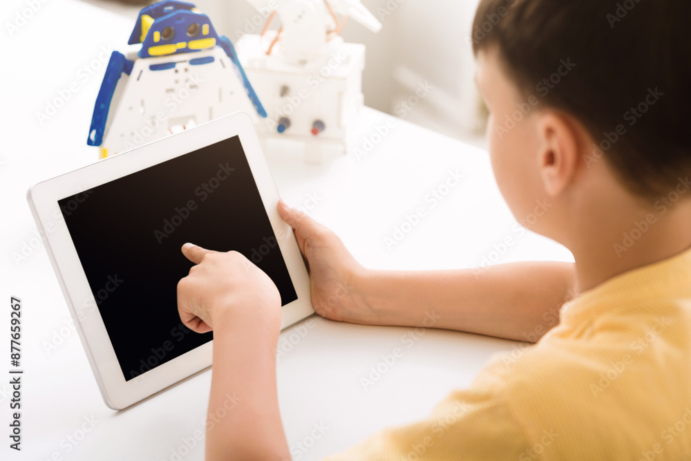 A young boy, sitting at a table, uses a tablet with his right hand and points at the screen. The screen is black. There is a partially constructed robot in the background. A young boy, sitting at a table, uses a tablet with his right hand and points at the screen. The screen is black. There is a partially constructed robot in the background.