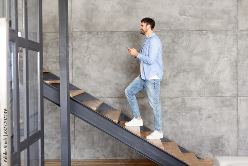 This image shows a man walking up a modern staircase in a home. The man is casually dressed in a blue shirt and jeans and is holding a phone in his hand. The stairs are made of wood © Prostock-studio