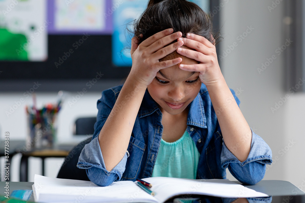 Stressed biracial schoolgirl sitting at desk in class, head in hands, staring at book, copy space