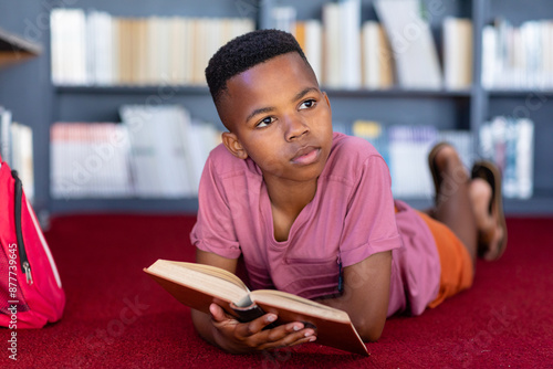 Thoughtful african american schoolboy lying on floor with book in library, looking away, copy space