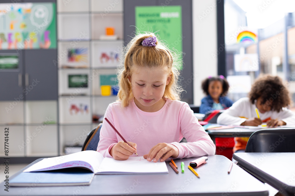Fototapeta premium Happy caucasian schoolgirl sitting at desk and writing in notebook in school classroom