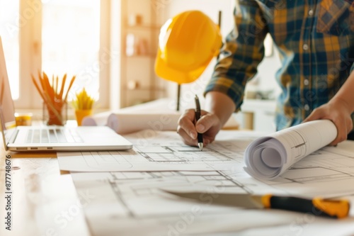 Architect at work with architectural plans and a laptop. A hard hat and stationery are on the table, indicating a construction environment.