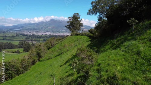Drone flight on the sunny side of a green hill with the view on the El corazón volcano in Ecuador on a sunny day with blue sky, copy space