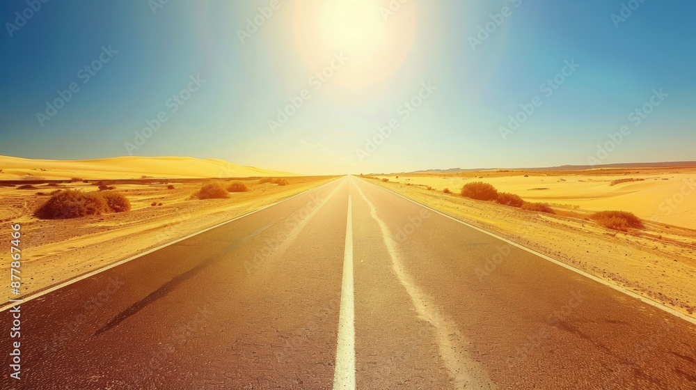 Naklejka premium A highway with a dashed white line down the center, stretching into the horizon through a desert landscape under a blazing hot sun. The road is flanked by sand dunes and sparse desert vegetation, with