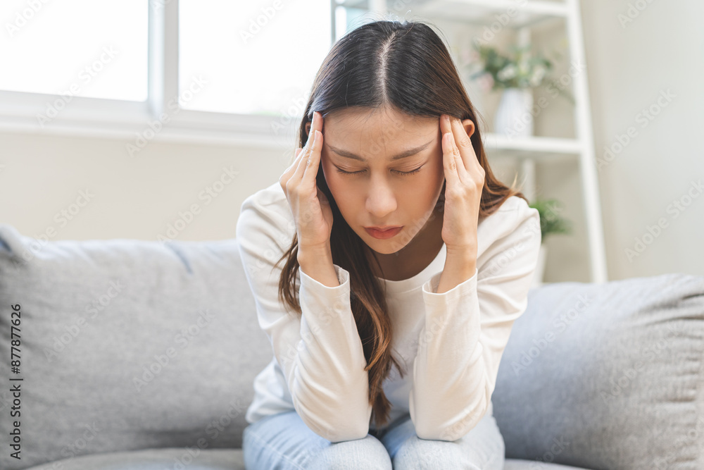 Dizzy asian young woman, girl headache or migraine pain suffering from vertigo while sitting on couch in living room at home, holding head with hand, health problem of brain or inner ear not balance.