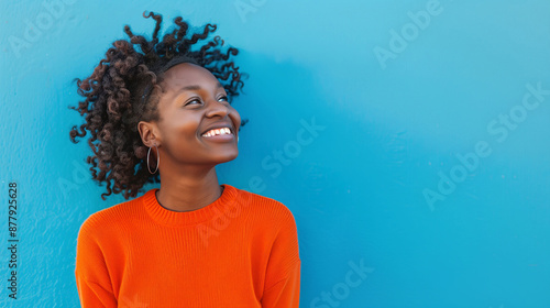 Joyful woman with curly hair smiling brightly against a vibrant blue background, wearing an orange sweater.