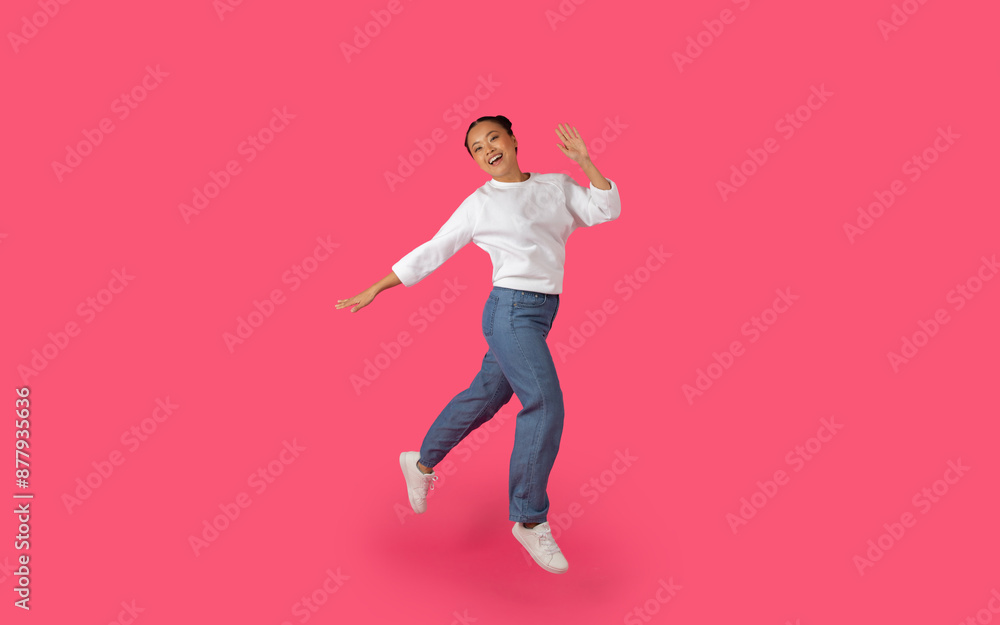 Asian young woman jumps and smiles in front of a bright pink background, her arms outstretched and her white shirt billowing in the air. Asian young woman jumps and smiles in front of a bright pink background, her arms outstretched and her white shirt billowing in the air.