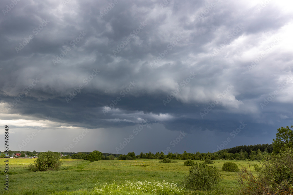 A large, ominous storm cloud looms over a peaceful spring meadow. Green grass and trees fill the foreground, while the dark sky above suggests a coming downpour. A large, ominous storm cloud looms over a peaceful spring meadow. Green grass and trees fill the foreground, while the dark sky above suggests a coming downpour.