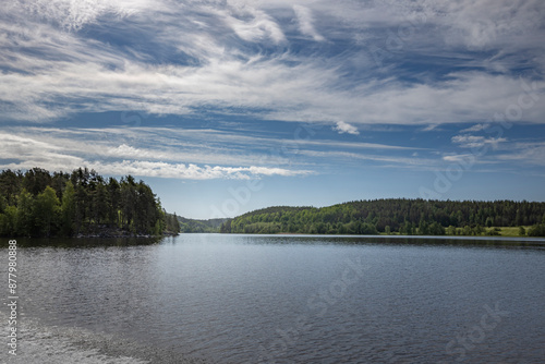 A serene view of a tranquil lake nestled within a lush forest, with a clear blue sky adorned with wispy clouds. The reflection of the sky creates a mirror-like surface on the water.