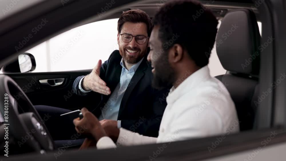 Discussing car features, options, and purchase details with friendly conversation. Caucasian car dealer sit inside vehicle with African American buyer. Discussing car features, options, and purchase details with friendly conversation. Caucasian car dealer sit inside vehicle with African American buyer.