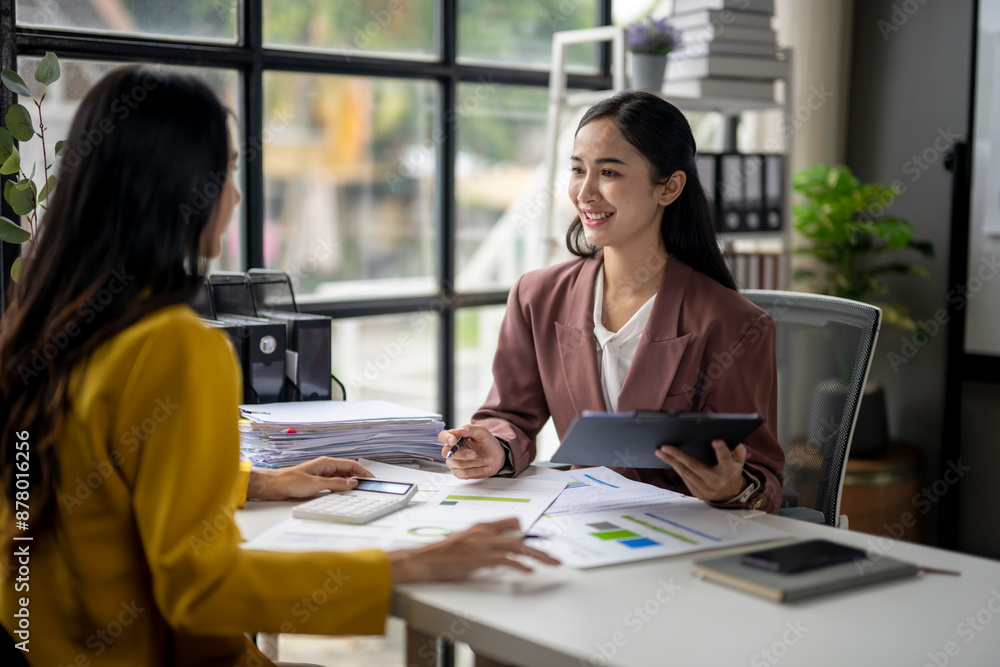 Two women are sitting at a desk, one of them holding a tablet Two women are sitting at a desk, one of them holding a tablet