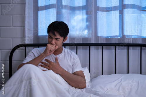 Φωτογραφία Asian unwell man sitting in bed at night, coughing and holding his chest indicat
