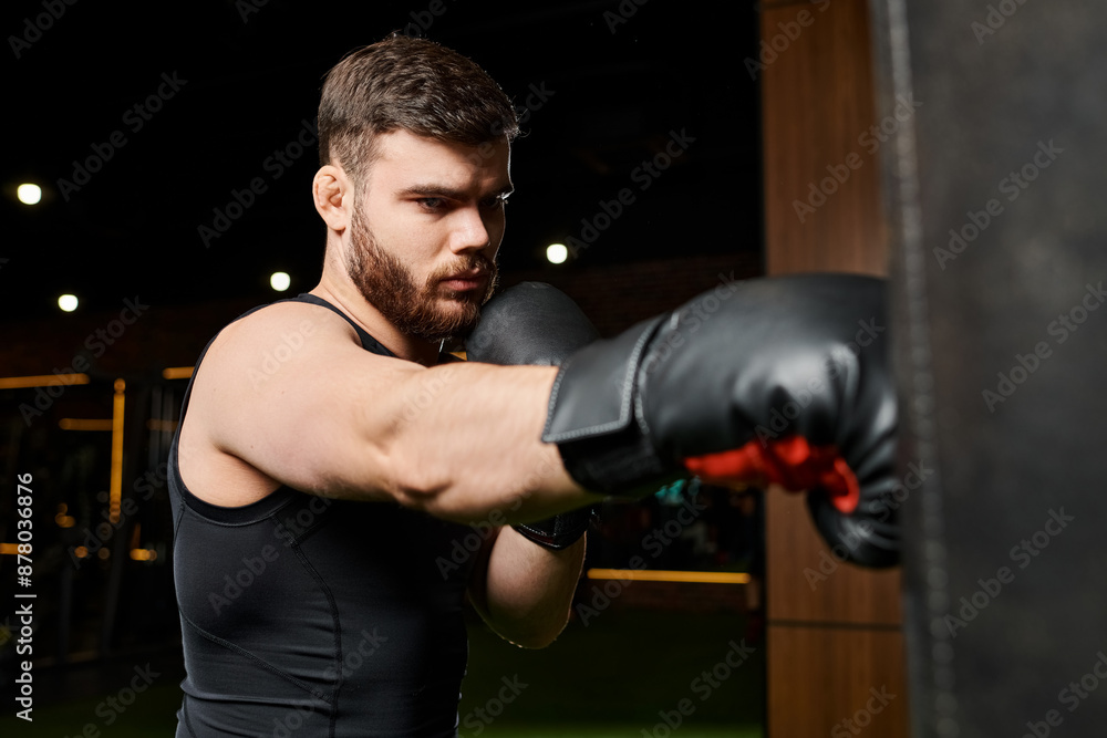 A handsome man with a beard wearing boxing gloves, throwing punches at a punching bag in a gym.