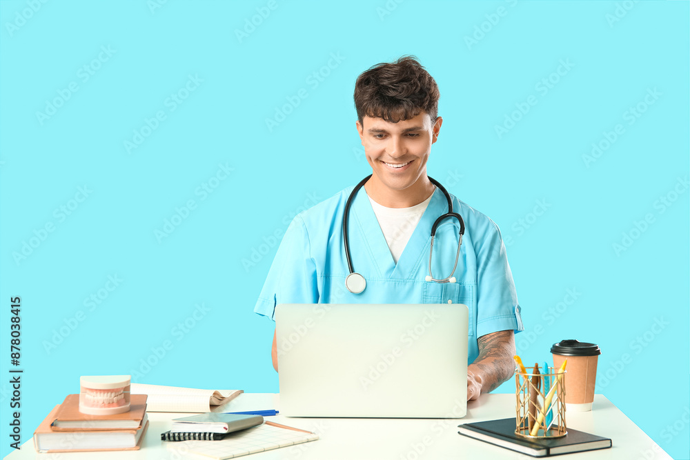 Male medical student with modern laptop, paper cup of coffee and jaw model at table on blue background