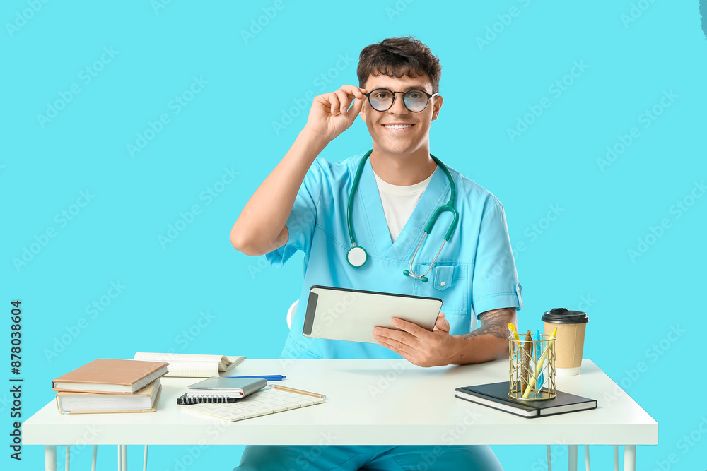 Male medical student with modern tablet computer, notebooks and paper cup of coffee at table on blue background Male medical student with modern tablet computer, notebooks and paper cup of coffee at table on blue background