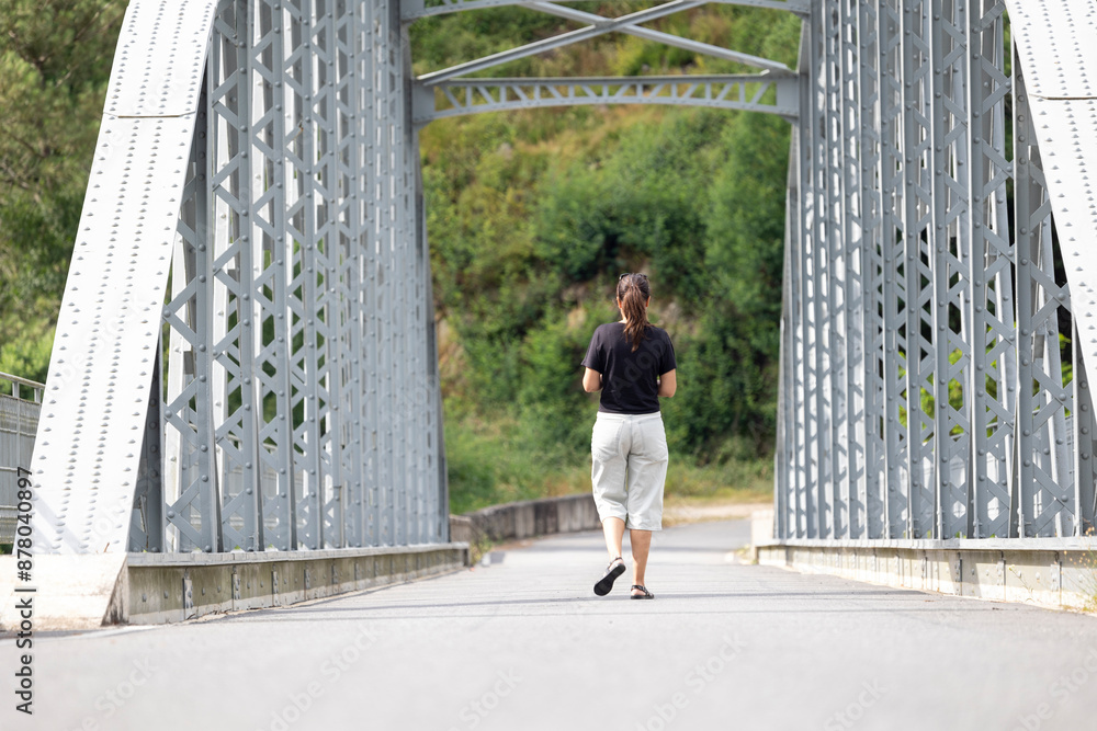 Ταπετσαρία Woman walking away on a sturdy, grey metal bridge surrounded by green trees and bushy vegetation, indicating a peaceful and serene outdoor environment
