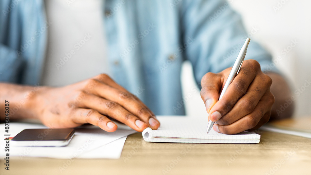 A person sits at a desk writing in a notebook with a pen. Their left hand is resting on a phone on the table. A person sits at a desk writing in a notebook with a pen. Their left hand is resting on a phone on the table.
