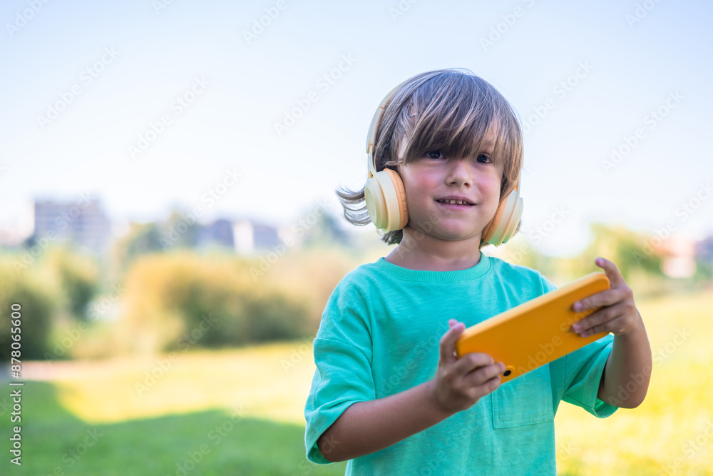 Portrait Of An Adorable 4 Years Old Boy Using Headphones And Smartphone, Stands In A Cute Outdoor Park.