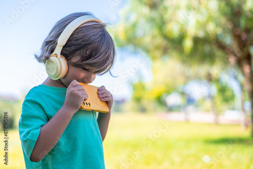 Portrait Of 4 Years Old Boy Using Headphones And Smartphone. Child Using A Tablet And Listening To Music In The Park photo