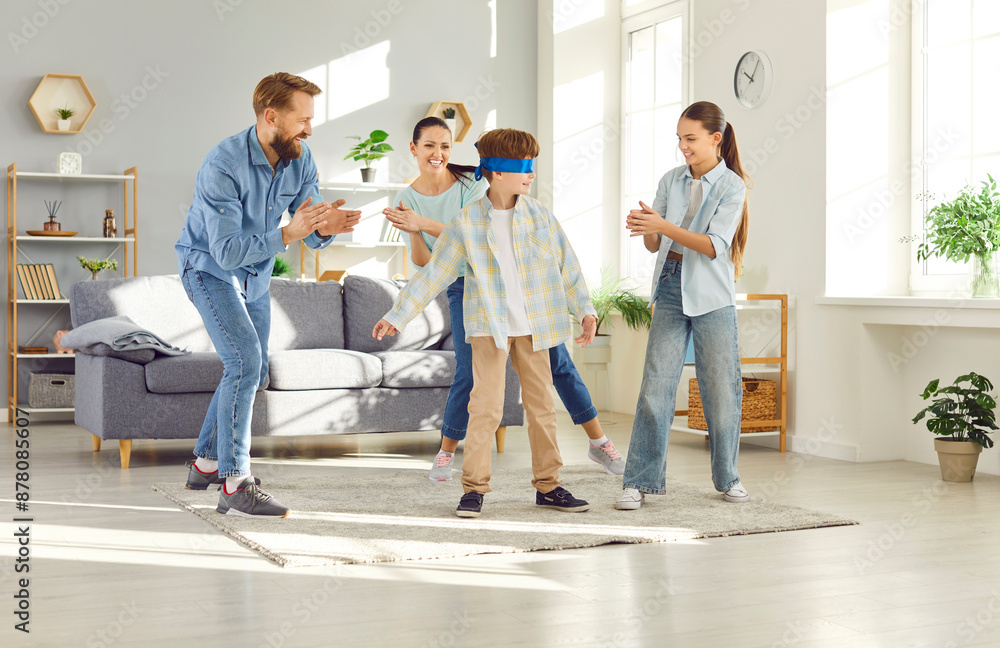 Cheerful mom and dad together with their children play game blind man buff in their home. In living room, blindfolded boy tries to catch other players while they clap clap to give audio clues. Cheerful mom and dad together with their children play game blind man buff in their home. In living room, blindfolded boy tries to catch other players while they clap clap to give audio clues.