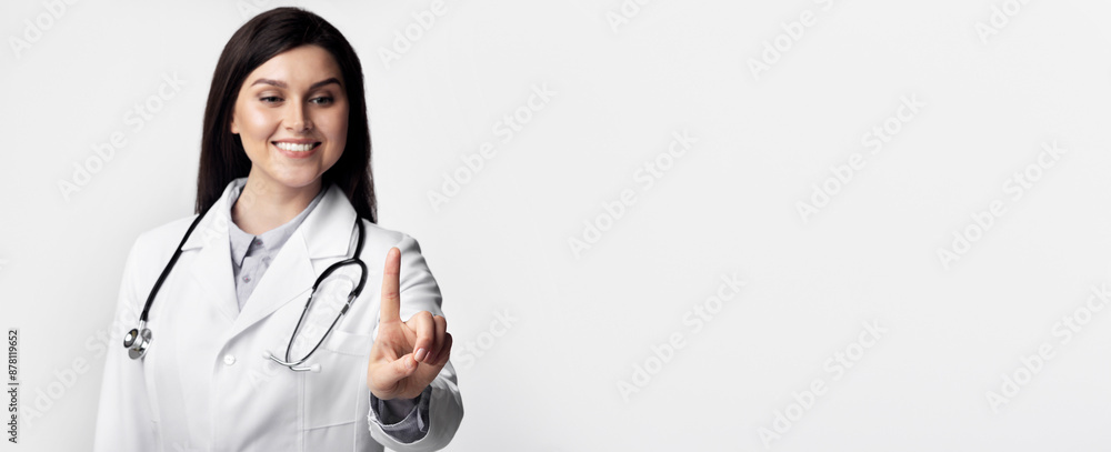 A young female doctor, wearing a white lab coat and stethoscope, smiles and points at a blank white background.