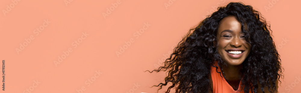 A smiling black woman with long, curly black hair stands against a peach-colored background. She is wearing a bright orange shirt and is looking directly at the camera. A smiling black woman with long, curly black hair stands against a peach-colored background. She is wearing a bright orange shirt and is looking directly at the camera.