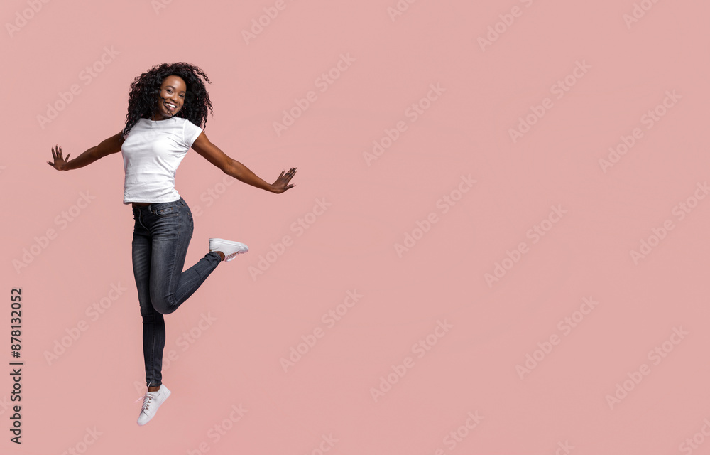 A young Black woman with curly hair is jumping in front of a pink background. She is wearing a white t-shirt and blue jeans. She is smiling and her arms are outstretched. A young Black woman with curly hair is jumping in front of a pink background. She is wearing a white t-shirt and blue jeans. She is smiling and her arms are outstretched.