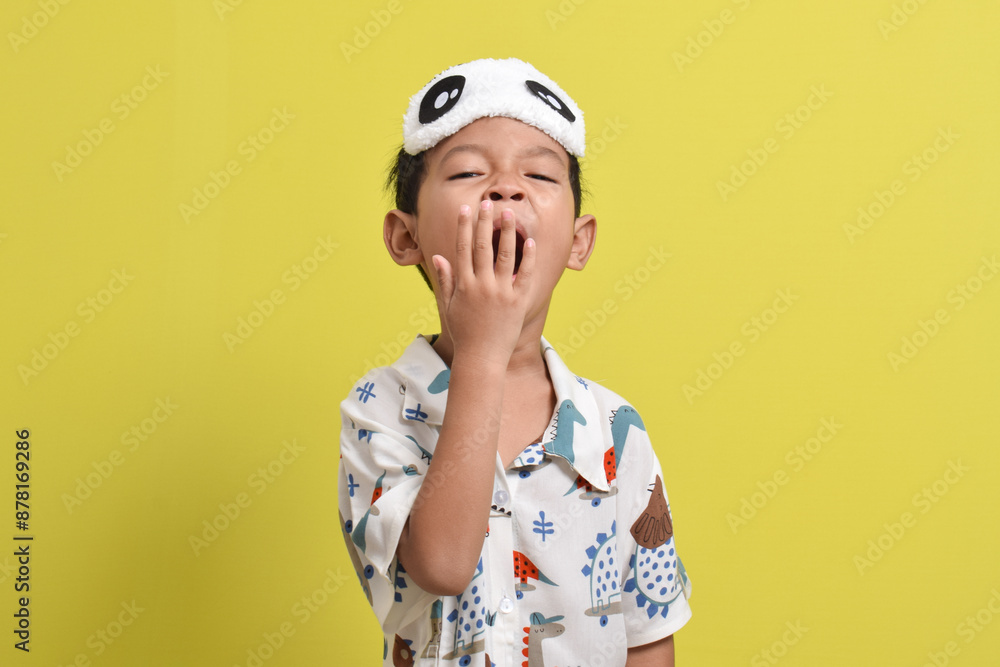 Portrait of Asian child sleepy and yawning, wearing pajamas and sleep mask isolated on yellow background. Child wants to sleep. Healthy sleep concept. Copy space Portrait of Asian child sleepy and yawning, wearing pajamas and sleep mask isolated on yellow background. Child wants to sleep. Healthy sleep concept. Copy space