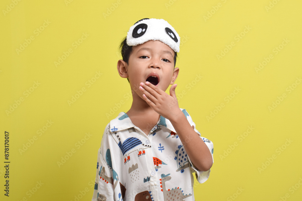 Portrait of Asian child sleepy and yawning, wearing pajamas and sleep mask isolated on yellow background. Child wants to sleep. Healthy sleep concept. Copy space Portrait of Asian child sleepy and yawning, wearing pajamas and sleep mask isolated on yellow background. Child wants to sleep. Healthy sleep concept. Copy space