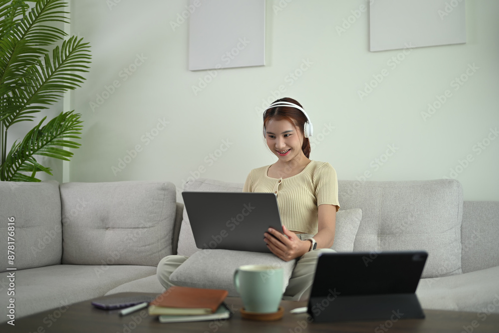 Charming young Asian woman in headphone using laptop on couch at home Charming young Asian woman in headphone using laptop on couch at home