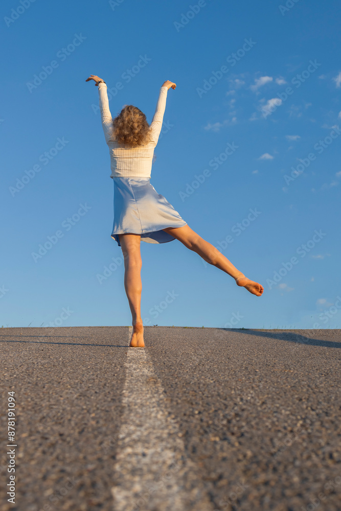 Obraz premium feminine, slender adult girl walks alone along the road to the blue sky and maintains her balance on the median. The concept of freedom, self-confidence and a bright future. feels great, festive