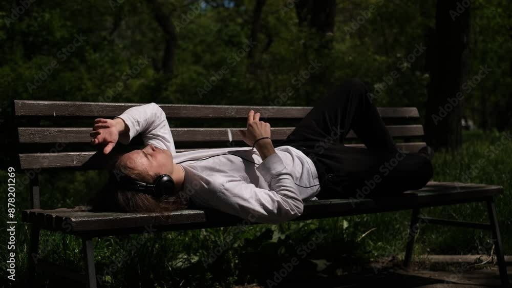 A young guy wearing headphones enjoys music while lying on a bench in the park. A young guy wearing headphones enjoys music while lying on a bench in the park.