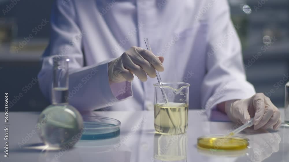 Video of a scientist doing an experiment with a yellow liquid, which contained in a glass beaker placed on a white laboratory table. The camera pans from left to right, space for text, front angle Video of a scientist doing an experiment with a yellow liquid, which contained in a glass beaker placed on a white laboratory table. The camera pans from left to right, space for text, front angle