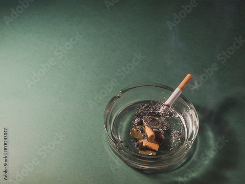 Fotografie Image of a cigarette burning in an ashtray with cigarette butts and coins