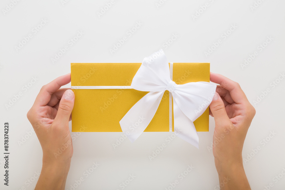 Young adult woman hands holding closed golden wedding envelope with white ribbon on light gray table background. Closeup. Point of view shot. Top down view.