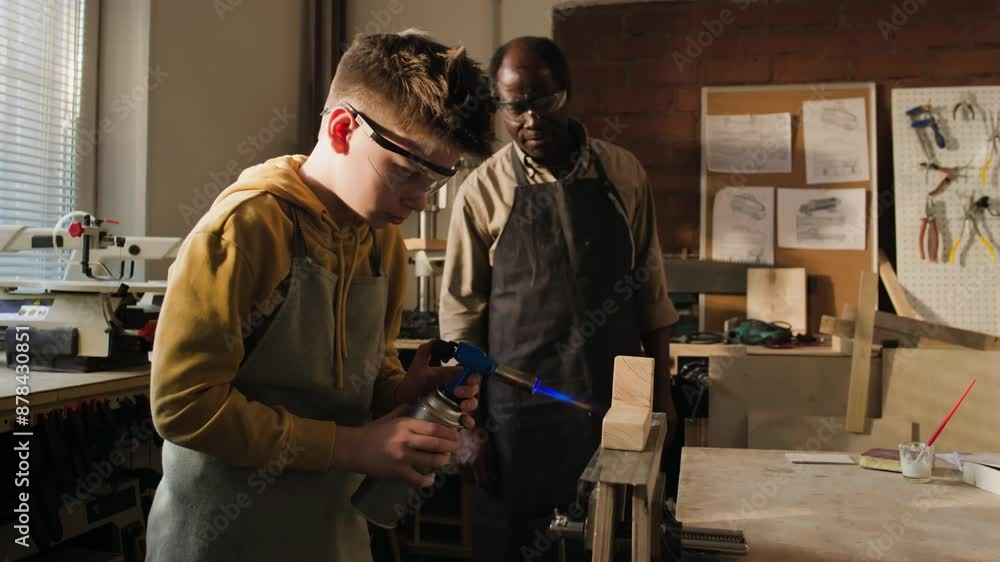 Little boy in safety glasses and apron using blow torch on wood under control of African American woodworking teacher in carpentry workshop Little boy in safety glasses and apron using blow torch on wood under control of African American woodworking teacher in carpentry workshop