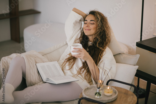 Papier peint Young woman reading book and drinking tea, sitting on sofa in a cozy living room