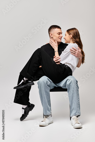 A young couple seated on a chair, sharing an intimate moment, surrounded by a grey studio background.