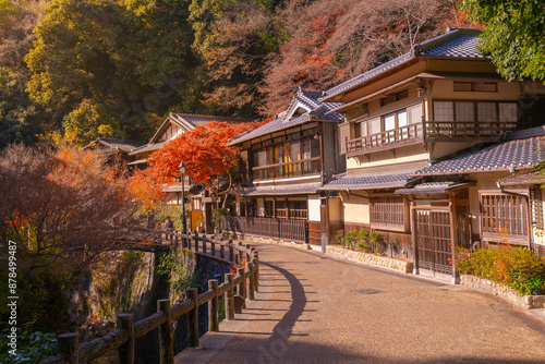 Traditional Japanese buildings line a picturesque path in Minoo Park, Kyoto, Japan. Vibrant autumn foliage creates a stunning canopy of red and orange leaves, highlighting the serene, historic atmosph photo