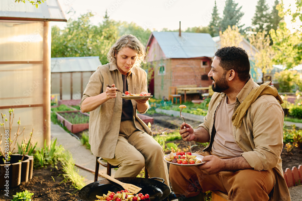 Naklejka premium Happy mature man with plate of vegetable stew looking at his wife eating cooked potatoes while both sitting by frying pan in the garden