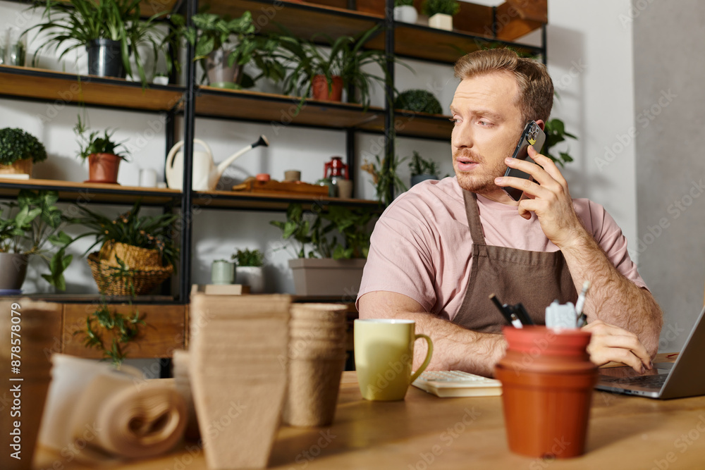 A man sits at a table in a plant shop, engrossed in conversation on his cell phone, embodying a small business owner in action. A man sits at a table in a plant shop, engrossed in conversation on his cell phone, embodying a small business owner in action.